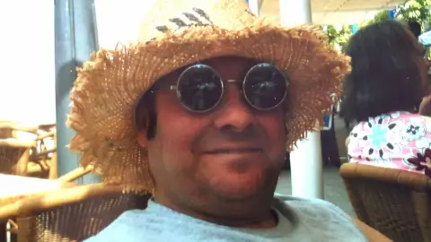 supplied Robert Newcombe smiles at the camera and is wearing a straw hat and a pair of sunglasses. He has a grey vest on.