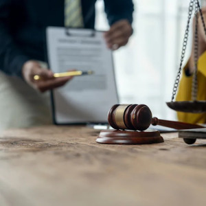 Person in business attire holding a clipboard with a document, while a gavel and scales of justice are in the foreground on a wooden table