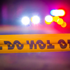 Blurred police lights behind yellow "Do Not Cross" tape, suggesting a crime scene at night