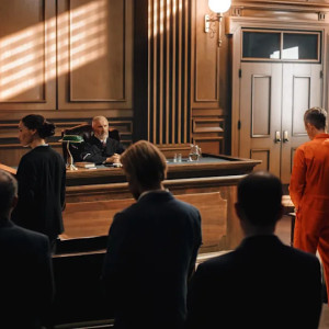 A courtroom scene shows a person in an orange jumpsuit with legal representatives present, standing before a judge